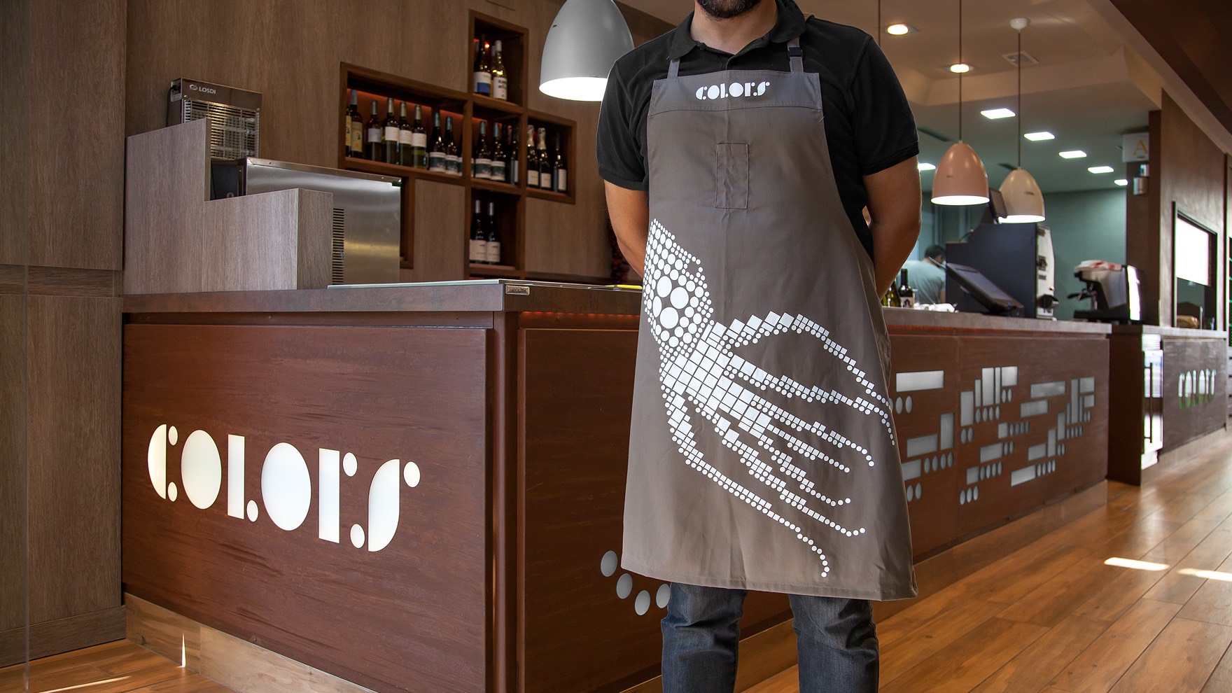 Camarero del restaurante COLORS con el delantal con la imagen corporativa aplicada y en el resto del restaurante tambi&eacute;n podemos ver la identidad aplicada.