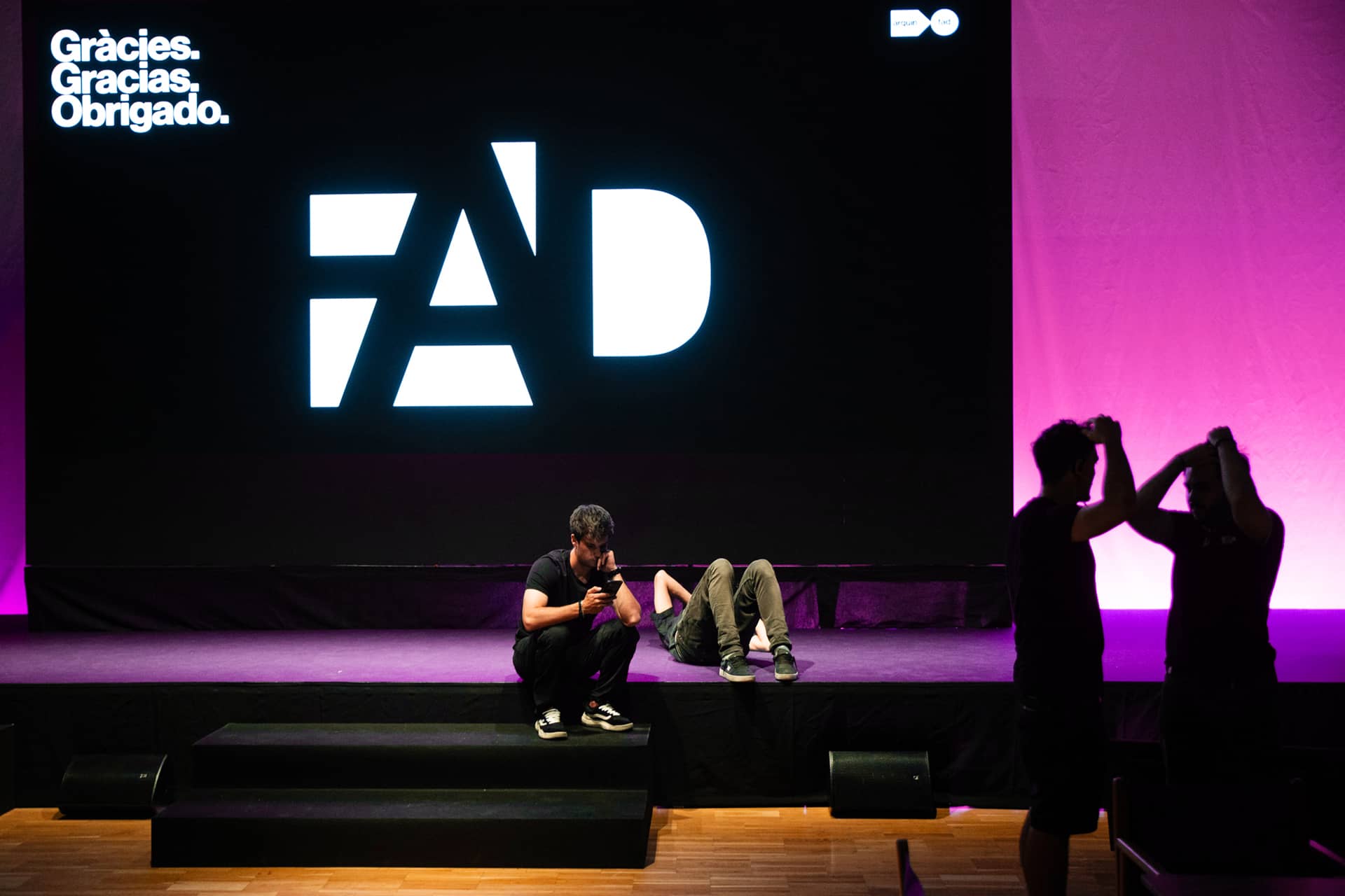 Dos chicos j&oacute;venes sentados en el escenario de la presentaci&oacute;n de la nueva identidad visual de los Premios FAD de Arquitectura e Interiorismo de 2024. Se muestra el logotipo geom&eacute;trico en plano y en blanco y negro de la nueva identidad visual de los Premios FAD de Arquitectura e Interiorismo de 2024.