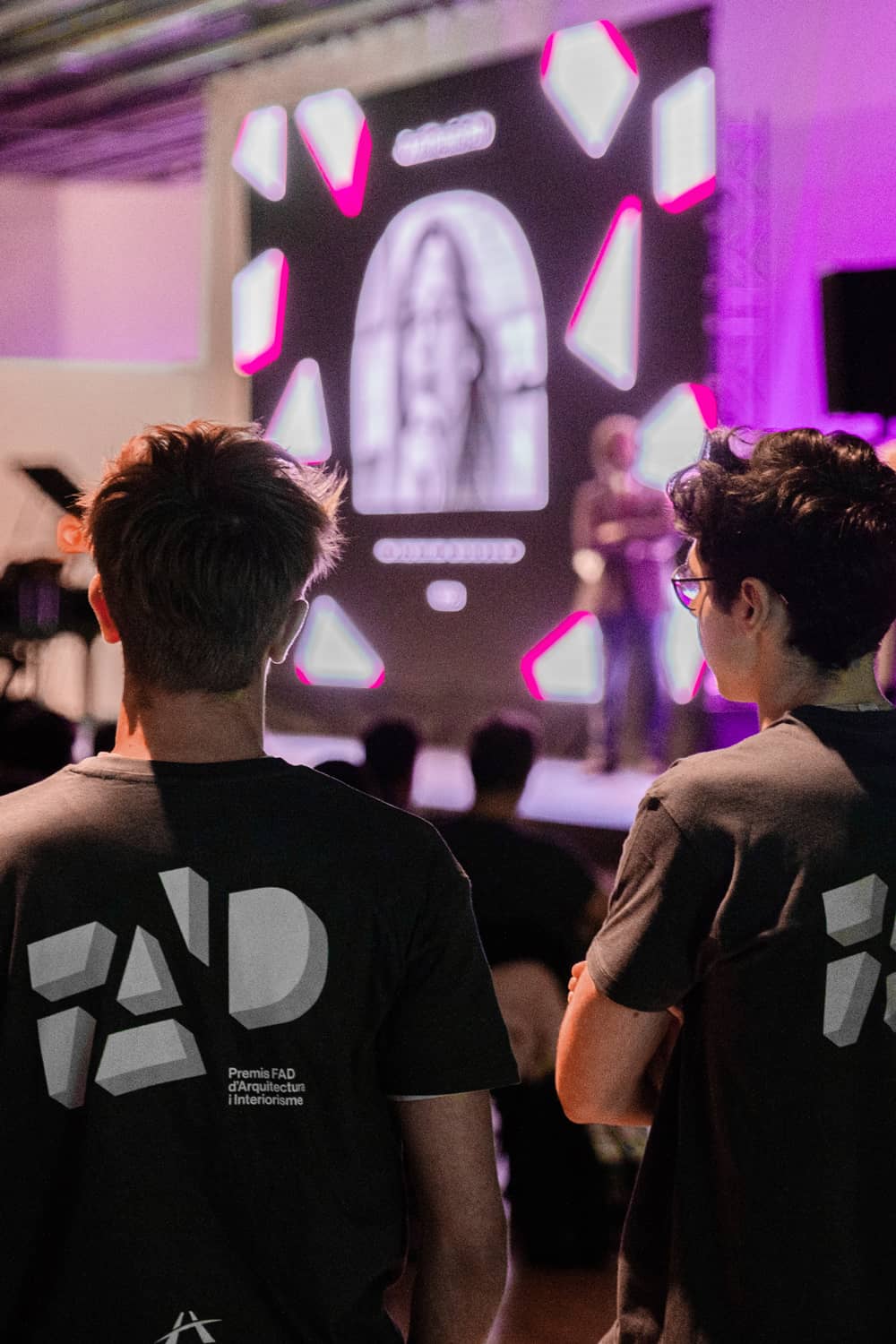 Dos camisetas cortas negras con, en la espalda, el dise&ntilde;o geom&eacute;trico del nuevo logotipo e identidad visual de los Premios FAD de Arquitectura e Interiorismo de 2024 en blanco y negro. Dos chicos j&oacute;venes mirando la presentaci&oacute;n de la nueva identidad de los Premios FAD 2024.