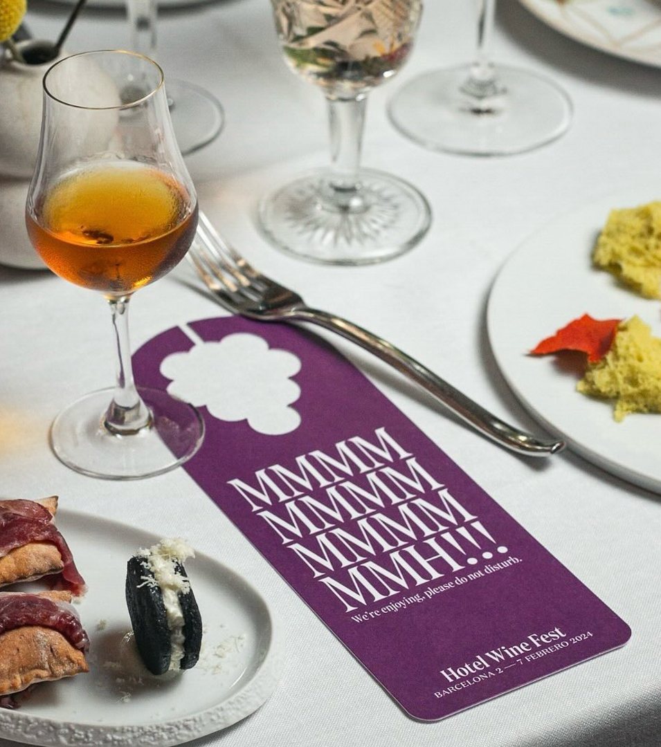 Un colgador de puerta de color morado sobre una mesa de restaurante, en el que pone Mmmmh!!! We&rsquo;re enjoying, please do not disturb, escrito con una tipograf&iacute;a serif moderna y con un agujero en el asa representando la silueta de un ramo de uvas. En la parte inferior, el logotipo tipogr&aacute;fico de Hotel Wine Fest y la fecha: Barcelona 2 &mdash; 7 Febrero 2024. Dise&ntilde;o que forma parte de la identidad y campa&ntilde;a del nuevo festival enogastron&oacute;mico, Hotel Wine Fest.