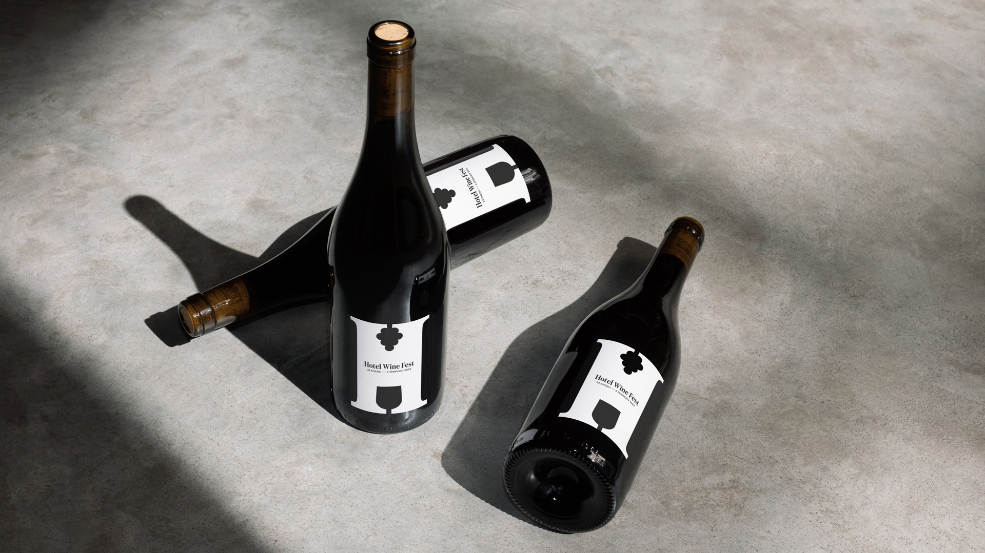 Dos botellas de vino tumbadas y una de pie sobre un suelo de piedra gris, las cuales contienen como etiqueta el icono de la letra &ldquo;H&rdquo; de Hotel Wine Fest en color blanco contrastado con el negro de la propia botella del vino y en una tipograf&iacute;a serif moderna.