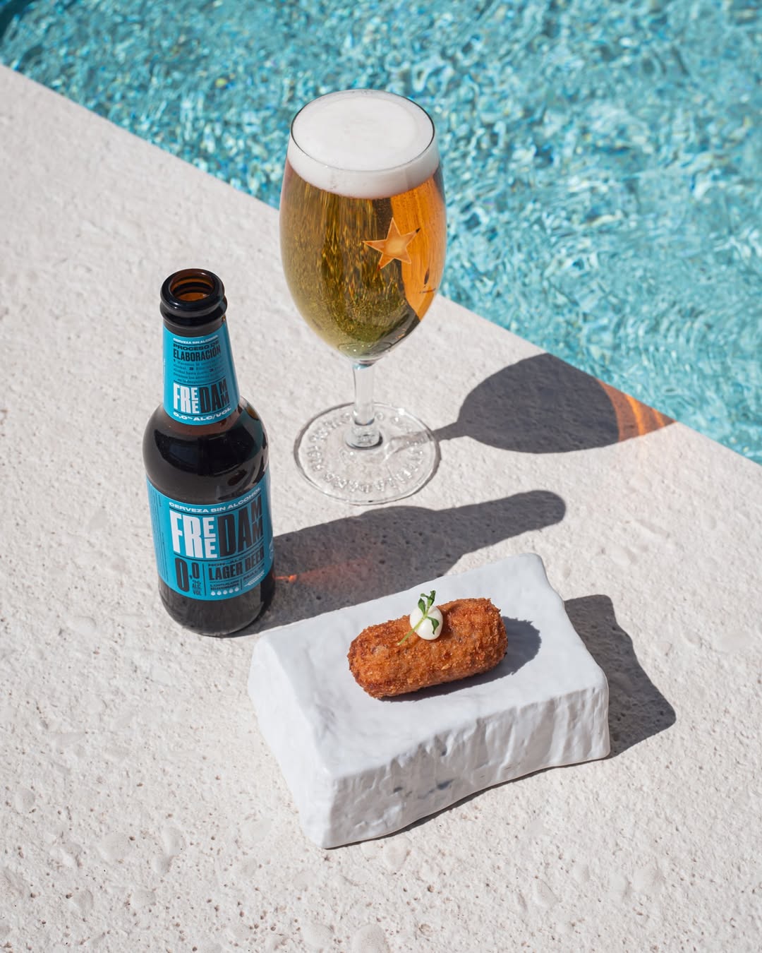 Cerveza botella Freedamm 0% alcohol con tapa espa&ntilde;ola junto a una piscina.